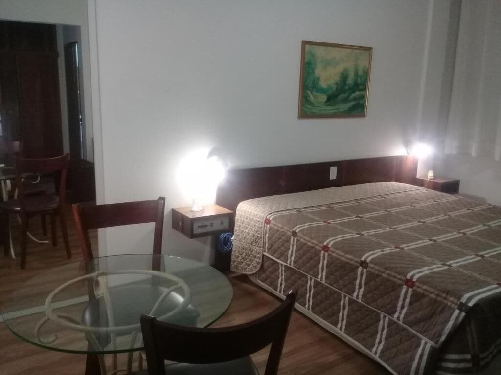 A hotel room with a bed, a glass table, a chair, a desk, and a nightstand with a lamp.