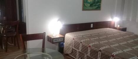A hotel room with a bed, a glass table, a chair, a desk, and a nightstand with a lamp.