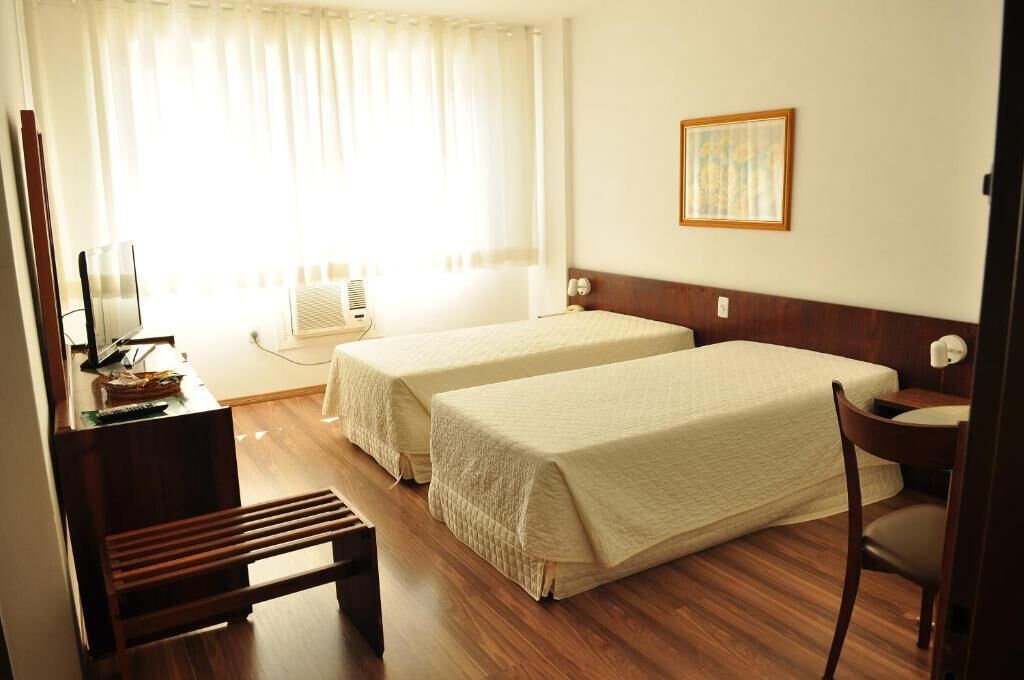 A hotel room with two single beds, a desk with a chair, a small table, a window with curtains, and a wall-mounted picture.