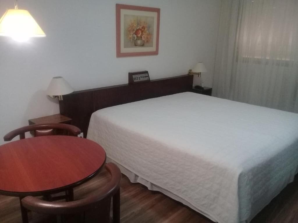 Habitación de hotel con una cama grande, una mesa redonda de madera, dos lámparas de noche y un cuadro en la pared.