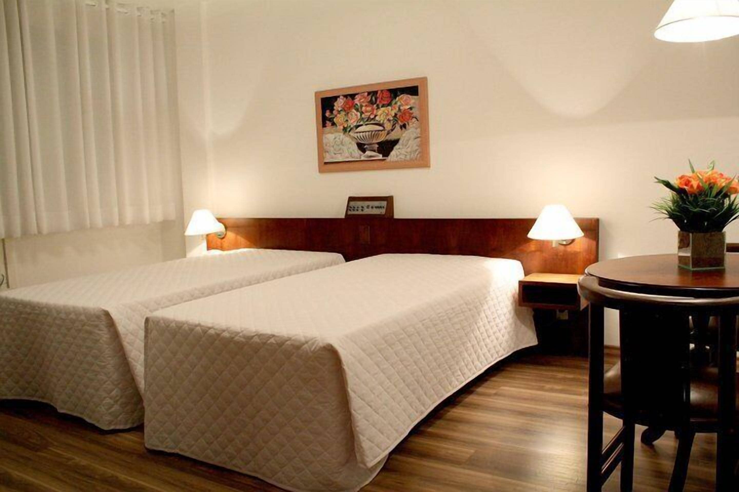 Een hotelkamer met twee bedden, een houten hoofdbord, bedlampen, een kleine tafel met een bloevas en een schilderij aan de muur.