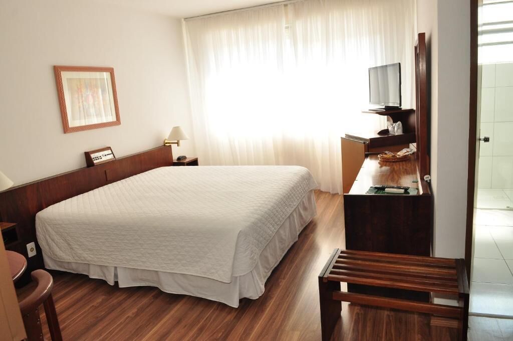 A hotel room with a large bed, a desk with a television, a chair, and a window with curtains.