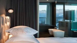 A hotel room with a bed, a nightstand, a lamp, a view of the mountains through the window, and a bathroom with a mirror and sink.
