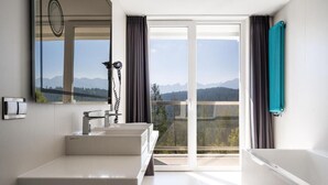 Un baño moderno con un espejo grande, dos lavamanos, una bañera y vistas a montañas a través de la puerta de vidrio.