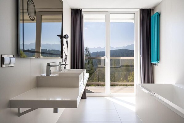 A modern bathroom with a large mirror, two sinks, a bathtub, and a view of mountains through the glass door.