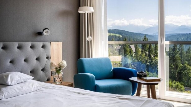 Habitación de hotel con una cama grande, un sillón azul, una mesita, una lámpara y vistas a las montañas a través de la ventana.