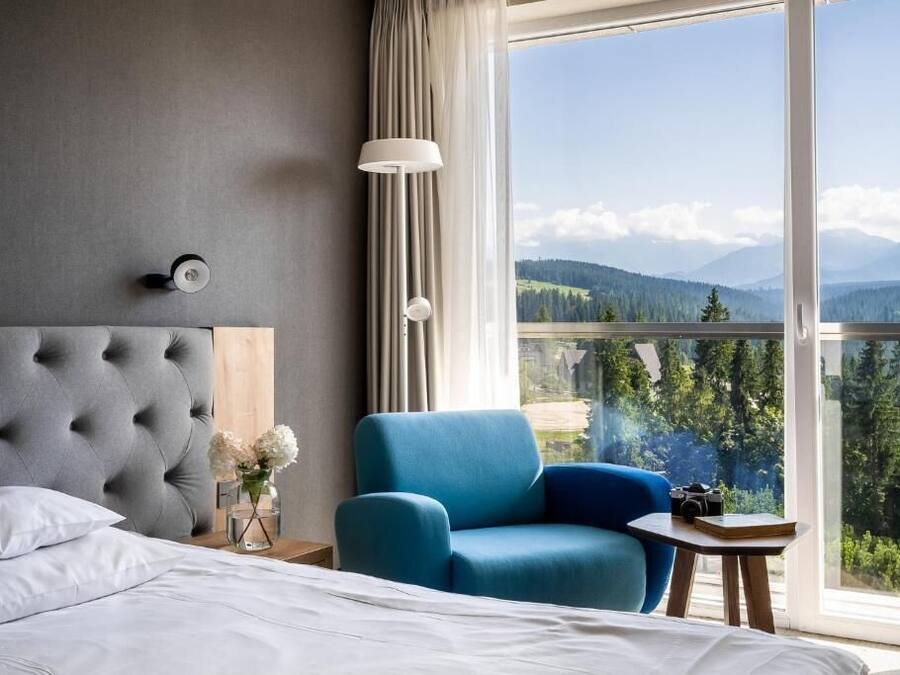 A hotel room with a large bed, a blue armchair, a small table, a lamp, and a view of mountains through the window.