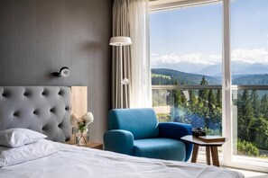 A hotel room with a large bed, a blue armchair, a small table, a lamp, and a view of mountains through the window.