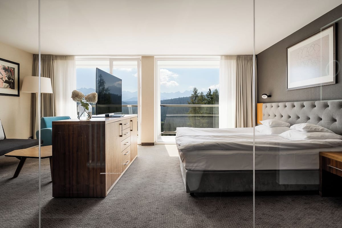 A modern hotel room with a large bed, a desk, a TV, and a view of mountains.