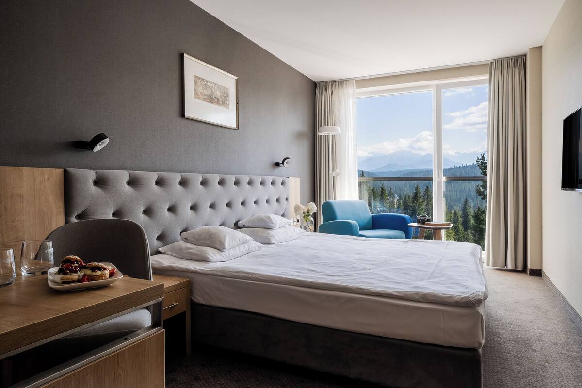 A hotel room with a large bed, a desk with a plate of food, a chair, a TV, and a view of mountains and a lake.