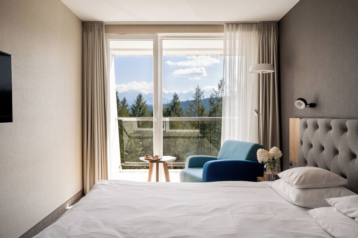 A hotel room with a large bed, a blue armchair, a small table with flowers, and a view of mountains through a large window.