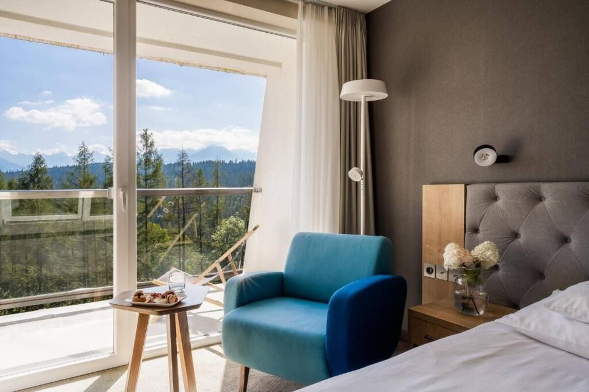 A hotel room with a bed, a chair, a small table with food, a vase with flowers, and a view of mountains through a large window.