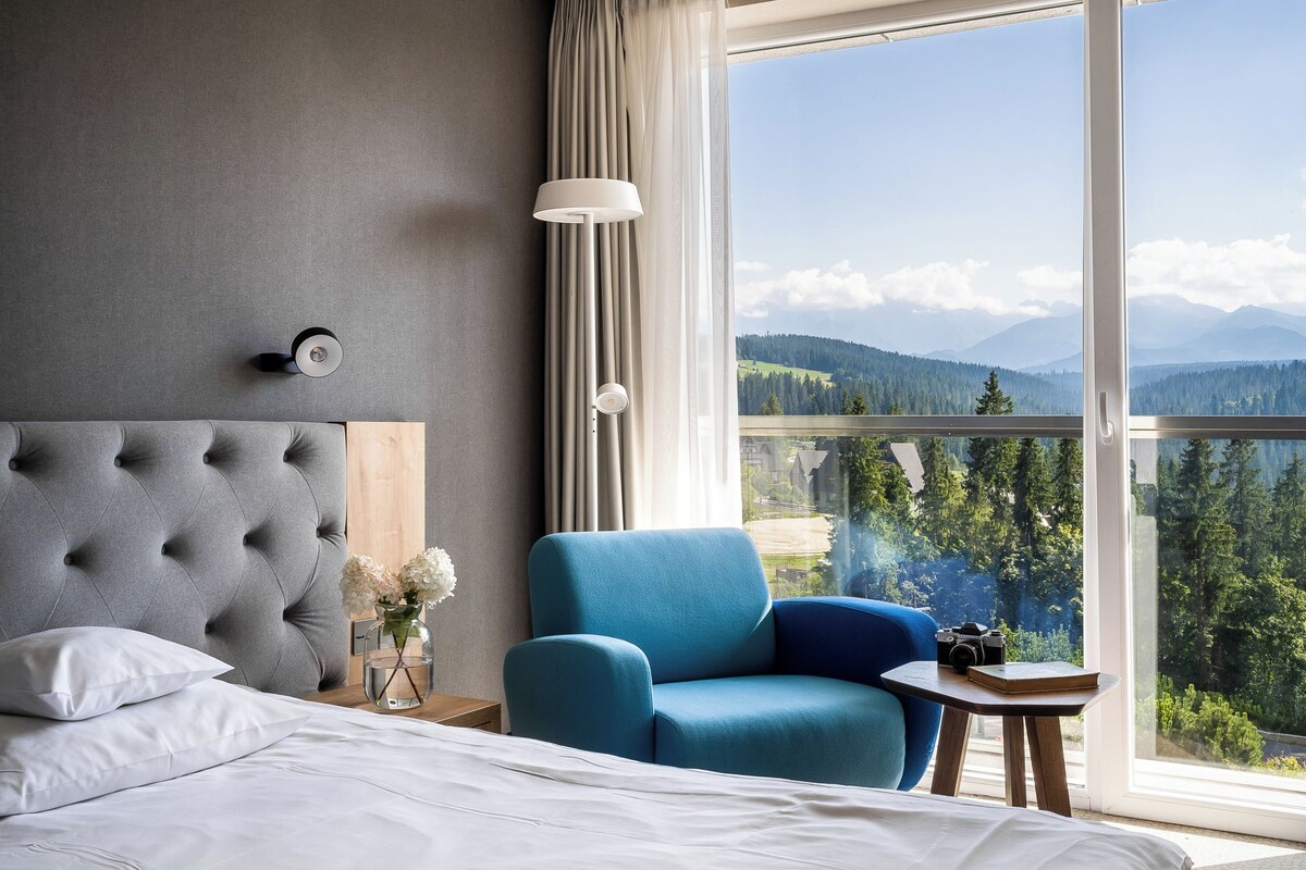 A hotel room with a bed, a blue armchair, a small table with a camera, and a view of mountains through large windows.