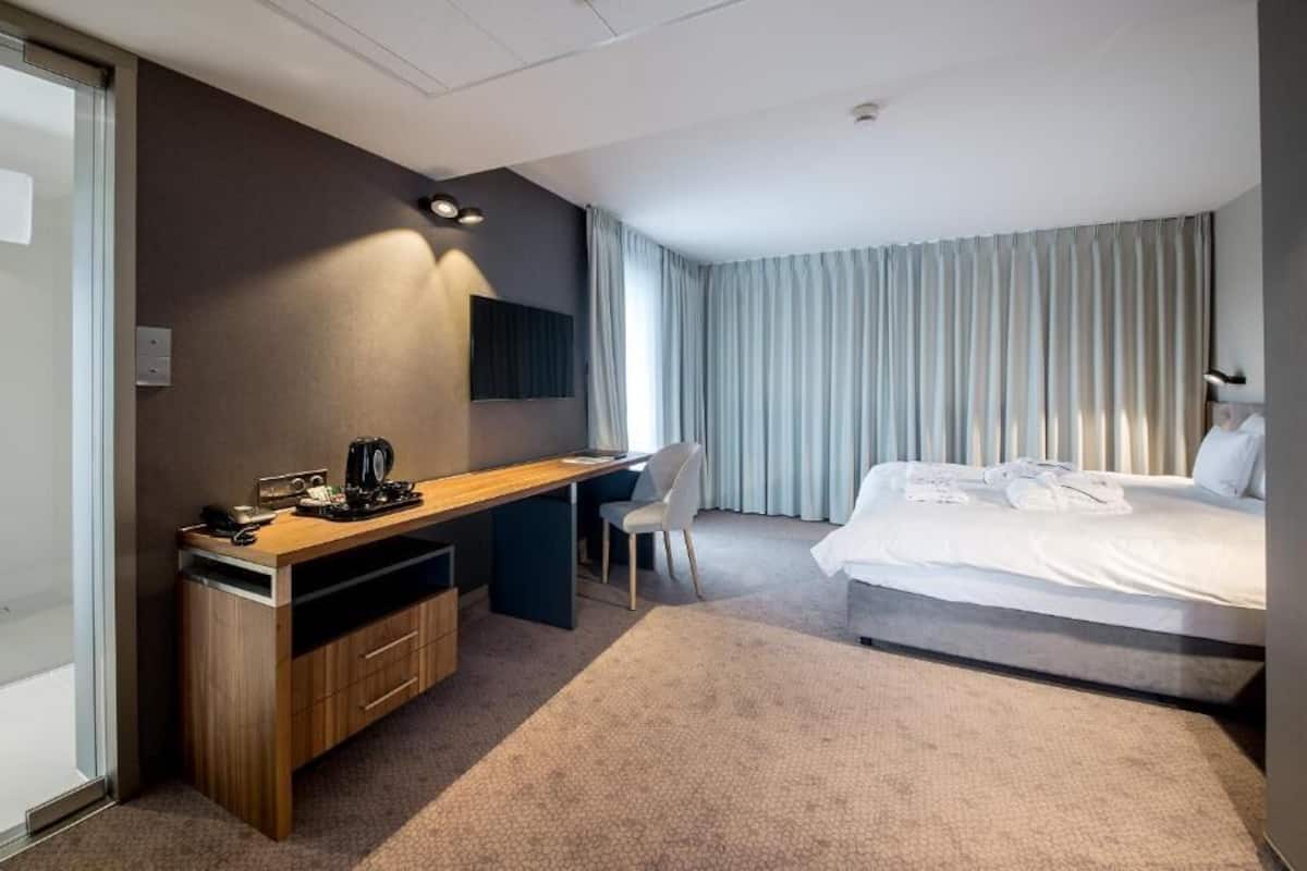 A hotel room with a bed, a desk, a chair, a TV, and a shower area.