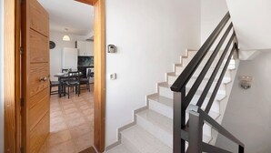 Uma escadaria com corrimão de madeira, uma área de cozinha com armários e um relógio, e um espaço de jantar com cadeiras e uma mesa.