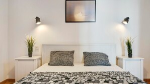 Quarto com cama, duas mesas de cabeceira com plantas, cabeceira e um quadro na parede.