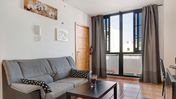 Uma sala de estar com um sofá cinza, uma mesa pequena, um vaso, uma porta de madeira e uma janela grande com cortinas.