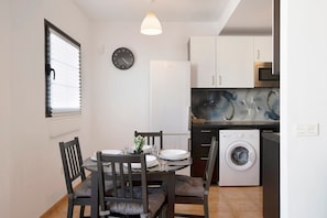 Une cuisine compacte équipée d’un réfrigérateur, d’un four à micro-ondes et d’un lave-linge, ainsi qu’une table à manger avec des chaises et une horloge au mur.