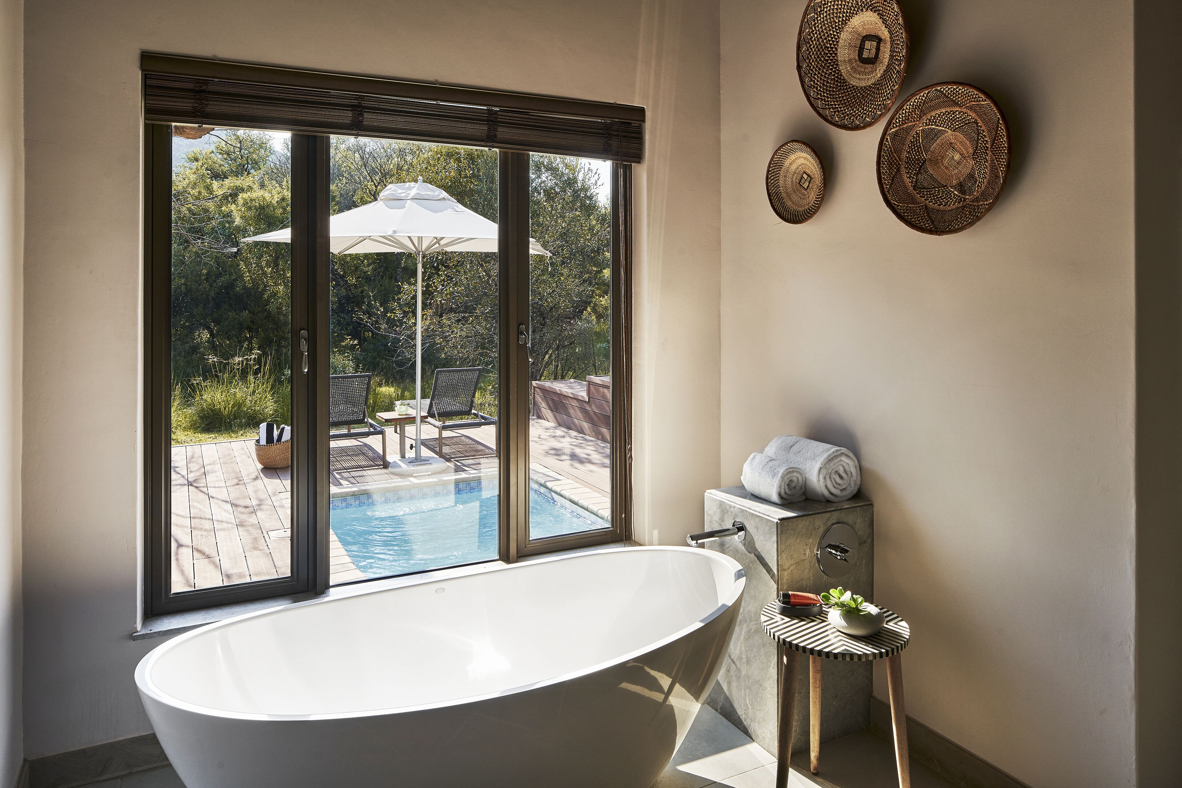 A modern bathroom with a freestanding bathtub, a small table with a plant, and a view of an outdoor pool area through large windows.
