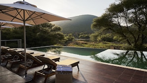 Una terraza de madera con sillones de descanso y una mesa, con vistas a una masa de agua tranquila y una colina boscosa.