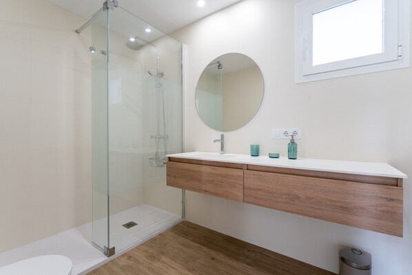 Un baño moderno con cabina de ducha de vidrio, un lavabo blanco sobre un mueble de madera y un espejo redondo.