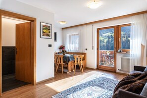 Una habitación con puerta de madera, un comedor con mesa y sillas, un ventanal con vistas a zonas verdes y una zona de estar acogedora con alfombra.