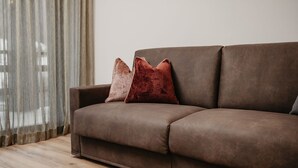 A brown sofa with two cushions, a wooden floor, and a sheer curtain.