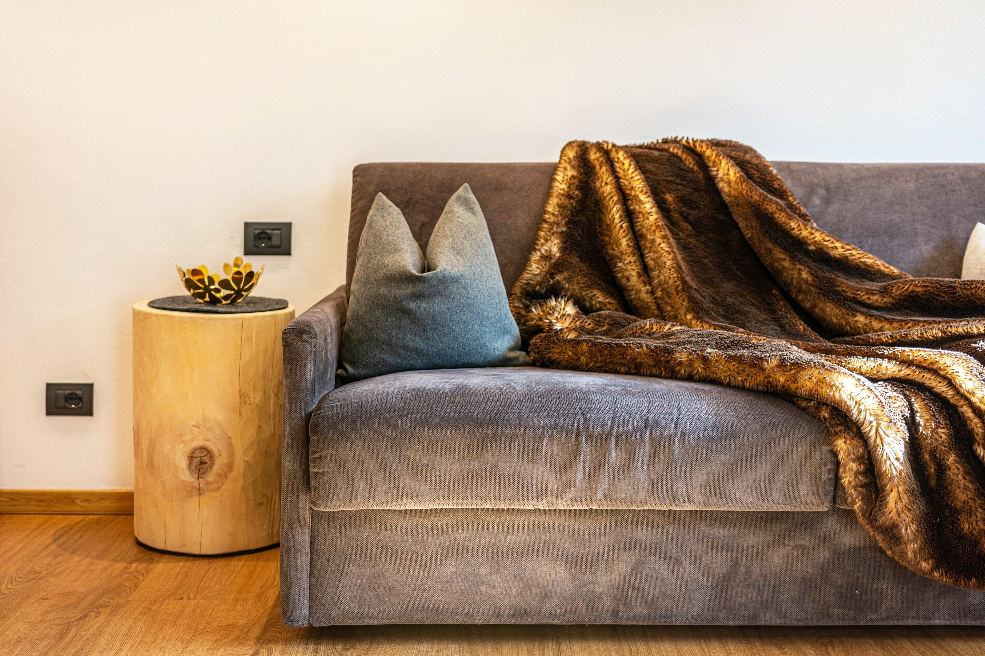 Un sofá gris con un plaid de piel sintética marrón, una almohada azul y una mesita de madera con un cuenco decorativo.