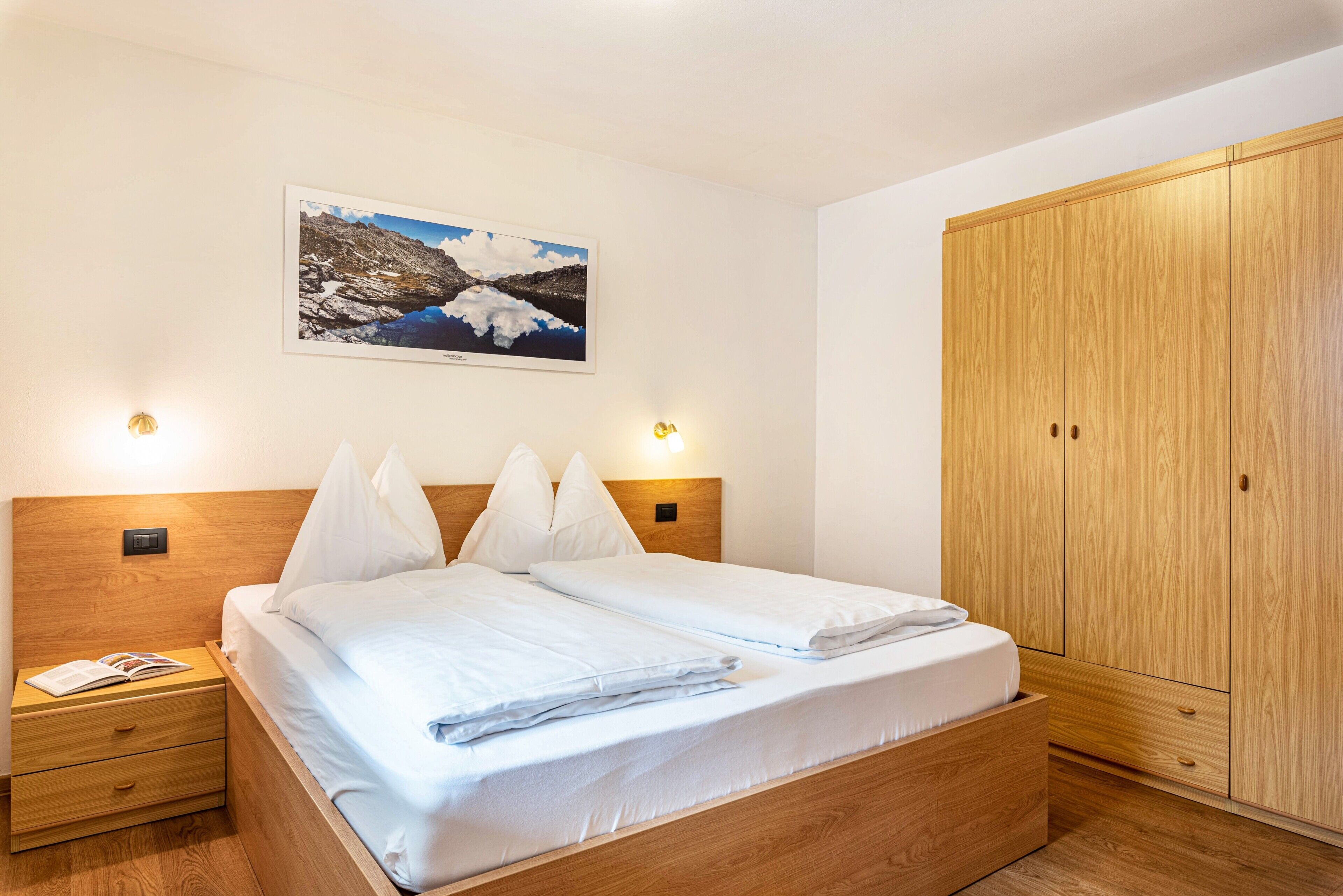 Une chambre à coucher avec un lit en bois, une table de chevet sur laquelle se trouve un livre, une armoire et un tableau accroché au mur.
