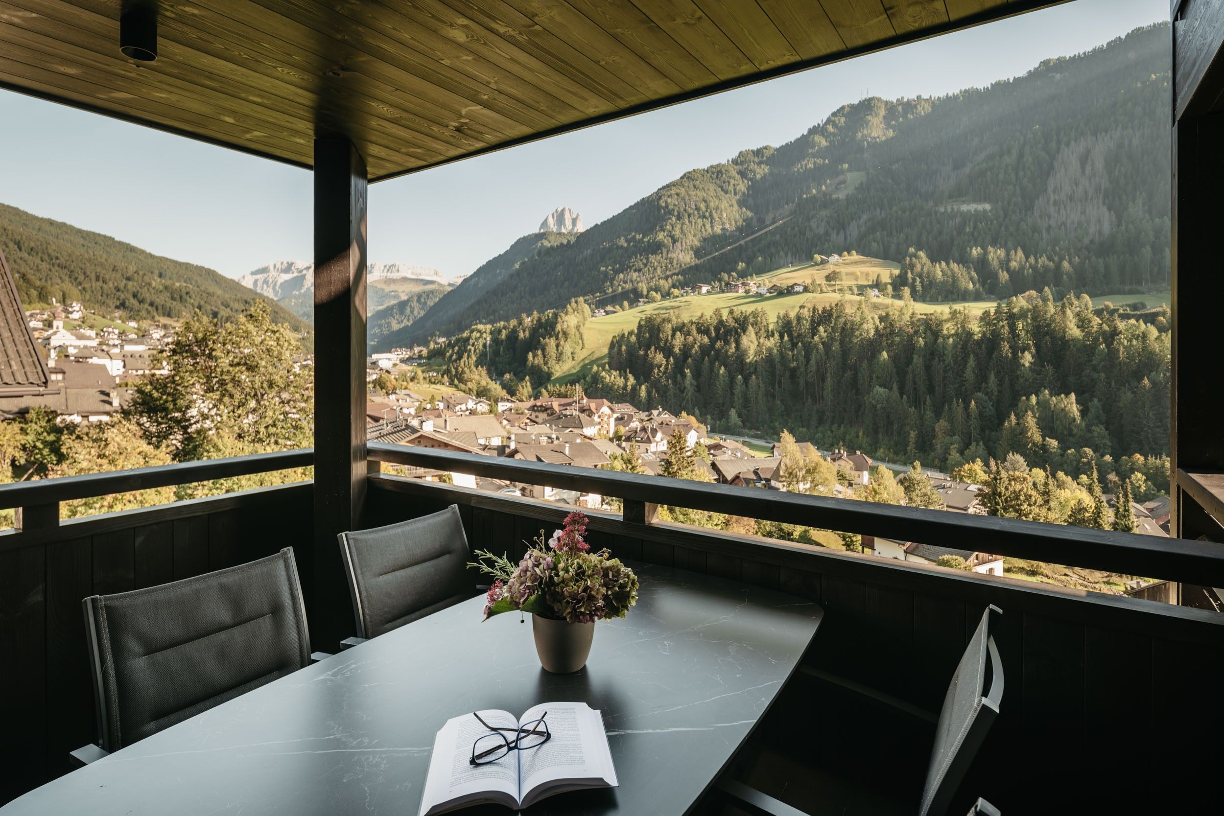A table with an open book and glasses, a potted plant, and a view of a valley with houses and mountains.