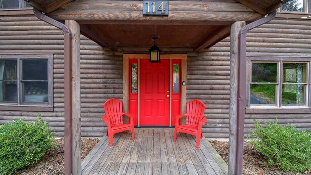 Une cabane en rondins avec une porte rouge, le numéro 114, deux fauteuils Adirondack rouges sur le porche et un petit jardin.