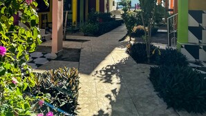Um pátio com caminho de azulejos, edifícios verdes e amarelos e flores cor-de-rosa.