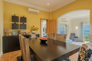 Un coin repas avec une table et des chaises en bois foncé, ainsi qu’un saladier. Des décorations sont accrochées aux murs et il y a un appareil de climatisation.