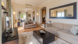 Un salon moderne avec un canapé, une table basse et un téléviseur. Un grand miroir orne le mur, et une fenêtre est agrémentée de rideaux.