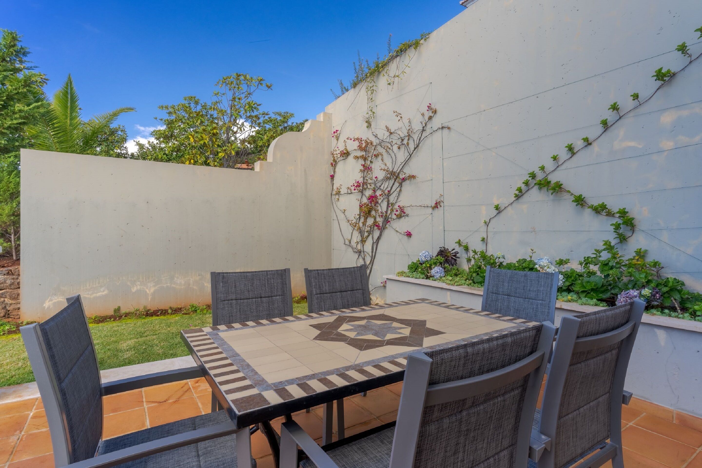 An outdoor patio with a table and chairs, surrounded by a white wall with climbing plants and flowers.