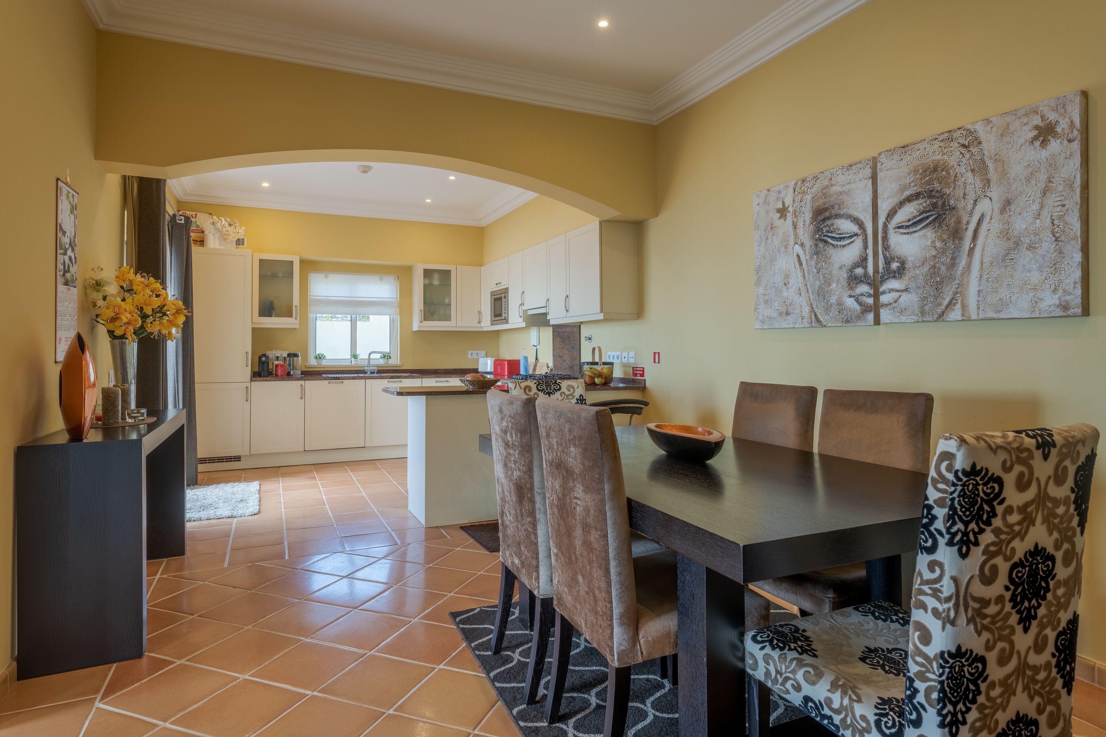 Un área de comedor con una mesa y sillas, una cocina al fondo y un cuadro de Buda en la pared.