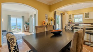 Un coin repas avec une table en bois foncé, des chaises de couleur beige et un saladier. De grandes fenêtres offrent une vue sur l’océan.