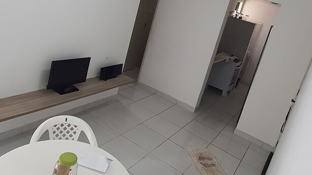 Una habitación pequeña y limpia con una mesa blanca, dos sillas, un banco blanco, un mueble blanco pequeño y una puerta que da a otra habitación.