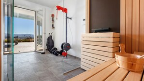 A modern gym with a glass door leading to a patio, weightlifting equipment, and a wooden sauna area. - Villa Ambretta with Private Swimming Pool, Garden and Barbecue (Campagnatico)