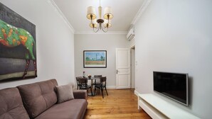 Una sala de estar con un sofá marrón, una mesa de comedor con sillas, un televisor de pantalla plana en la pared y una araña.
