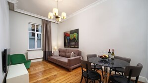 Un salon avec un canapé, une table à manger entourée de chaises, un fauteuil vert, un tableau accroché au mur et une fenêtre avec des rideaux.