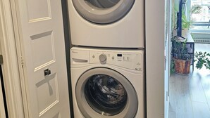 A compact, two-door washer and dryer unit in a hallway with a door on the left and a potted plant on the right.