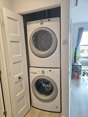 A compact, two-door washer and dryer unit in a hallway with a door on the left and a potted plant on the right.