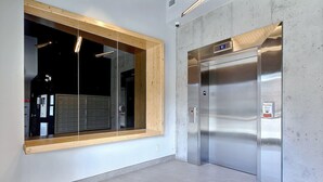 A modern elevator with a stainless steel finish and a digital display, adjacent to a large window with a wooden frame and a view of a building's interior.