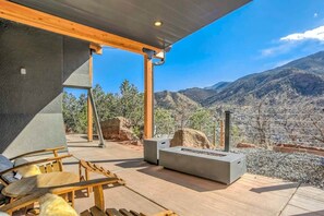 An outdoor patio with wooden furniture, a built-in grill, and a view of a mountainous landscape. - Luxury Retreat | Hot Tub, Sauna, Gym | Manitou (Manitou Springs)