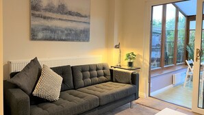 Een moderne woonkamer met een grijze bank, een schilderij, een lamp en uitzicht op het omliggende landschap door grote ramen.