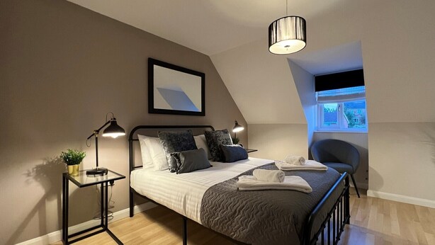 Een moderne slaapkamer met een bed, bedlampjes, een klein tafeltje met een plant en uitzicht buiten door een raam.