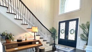 A grand staircase with a chandelier, a wooden bench, and a rug leading to a dark wooden door with decorative glass panels.