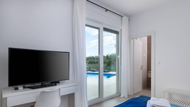 Une chambre d’hôtel moderne équipée d’un téléviseur à écran plat, d’un bureau, d’une chaise, d’un lit avec une literie bleue et blanche, et d’une porte-fenêtre coulissante donnant sur un espace piscine.
