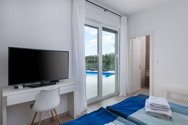 A modern hotel room with a flat-screen TV, a desk, a chair, a bed with blue and white bedding, and a sliding glass door leading to a pool area.